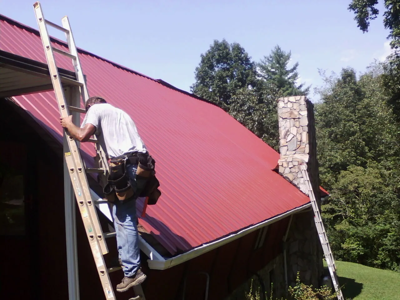 Roofer on a ladder beside a red metal roof and stone chimney — Soffit & Fascia Repair by Roofing Brothers in Wells River